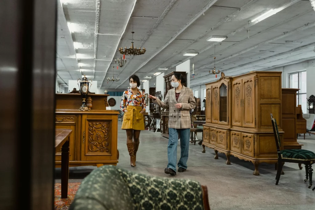  Deux femmes portant des masques de protection discutent tout en marchant dans un grand magasin de meubles anciens, entourées de buffets et armoires en bois sculpté. 