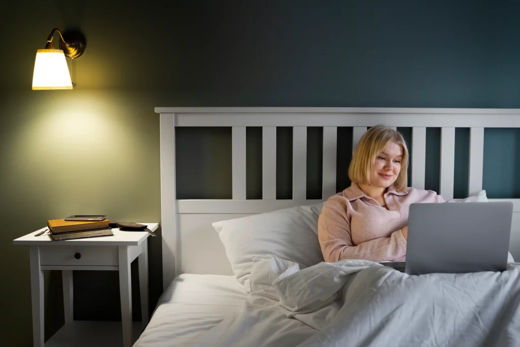 Femme avec un ordinateur portable sur son lit dans sa chambre éclairée grâce à une lampe murale